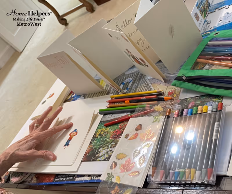 Craft table with cards, markers, and colored pencils; resident's hand visible.