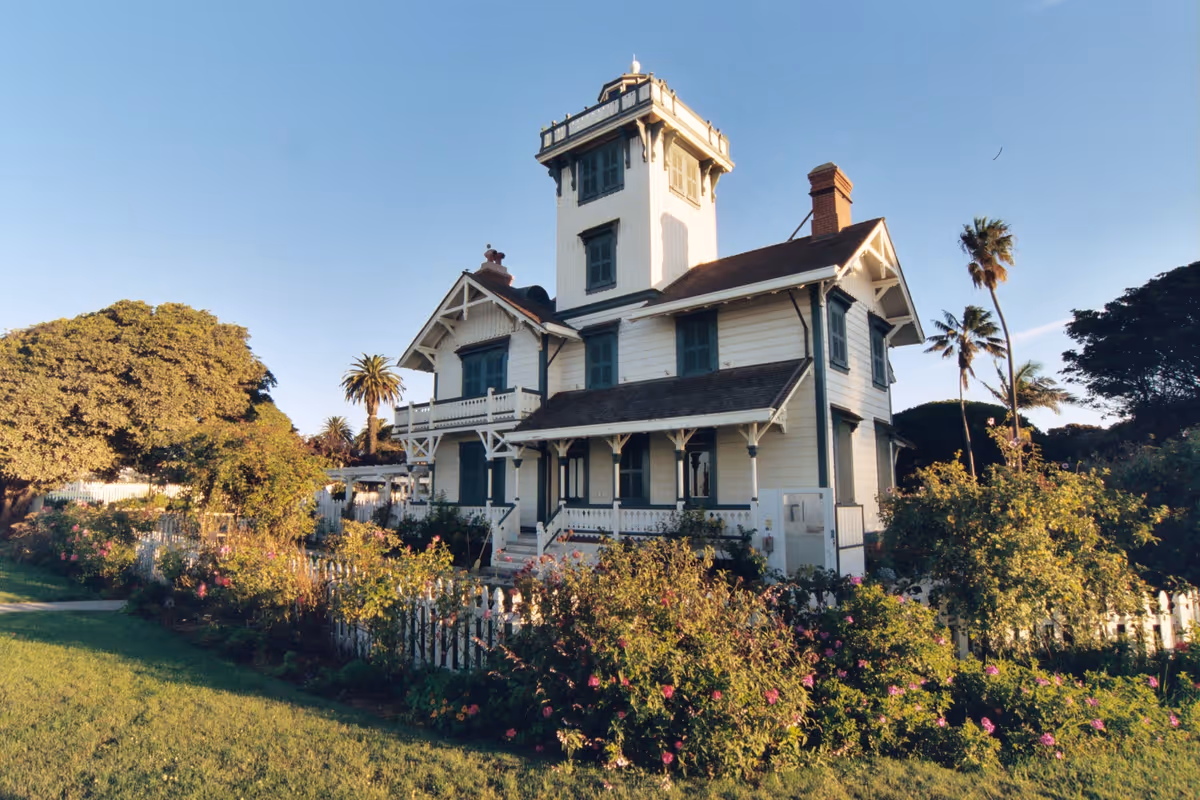 A large white Victorian-style house with a tower and wraparound porch in a garden.