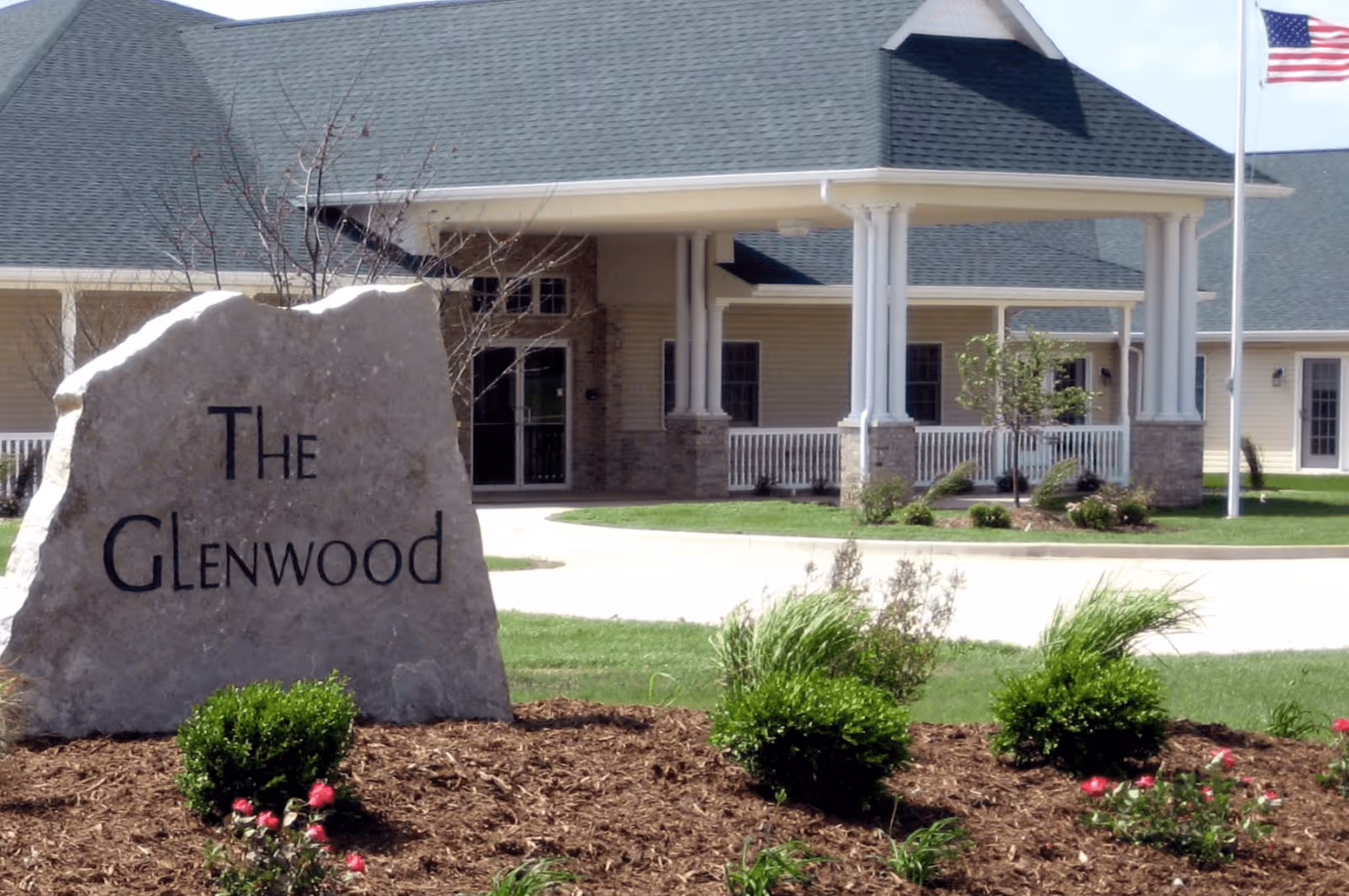 The entrance of The Glenwood Assisted Living of Mahomet facility with a large stone sign that reads 'The Glenwood' in front of a landscaped area with bushes and flowers. The building has a covered porch with white columns and an American flag on a flagpole to the right.