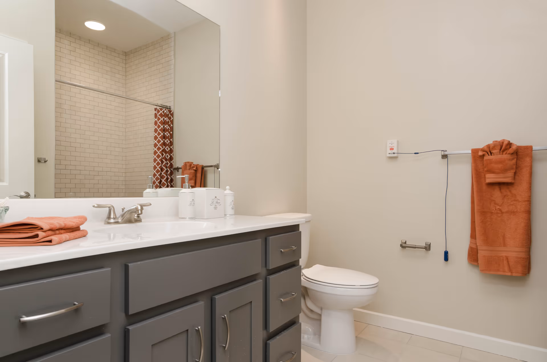 A clean and modern bathroom featuring a white toilet, a gray vanity with a white countertop, a large mirror above the sink, and a shower area with beige subway tiles and a patterned orange shower curtain. Orange towels are neatly folded on the countertop and hanging on a towel rack beside the toilet.