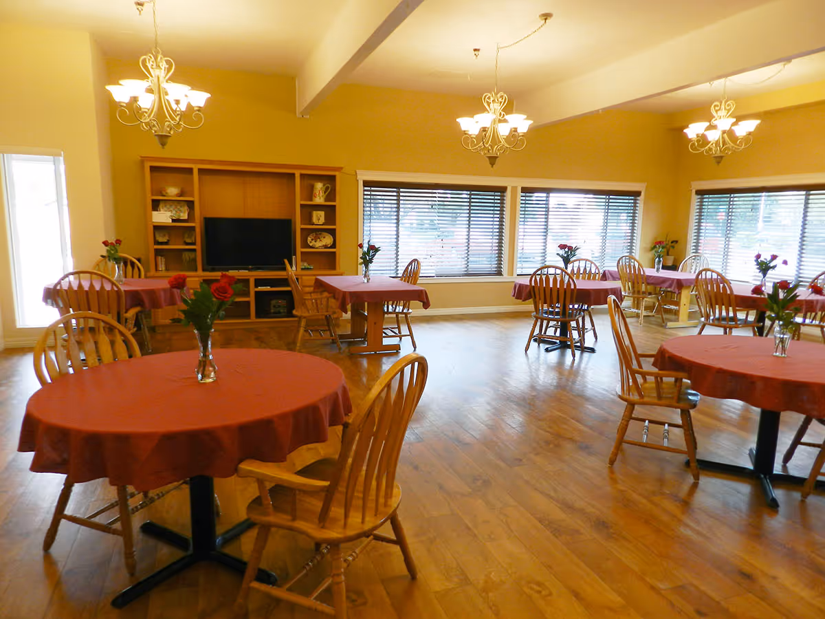 A dining room with several round tables covered with red tablecloths, each table having a vase with red flowers. Wooden chairs surround the tables. The room has large windows with blinds, wooden flooring, and three chandeliers hanging from the ceiling. A wooden entertainment unit with a TV and decorative items is visible against one wall.