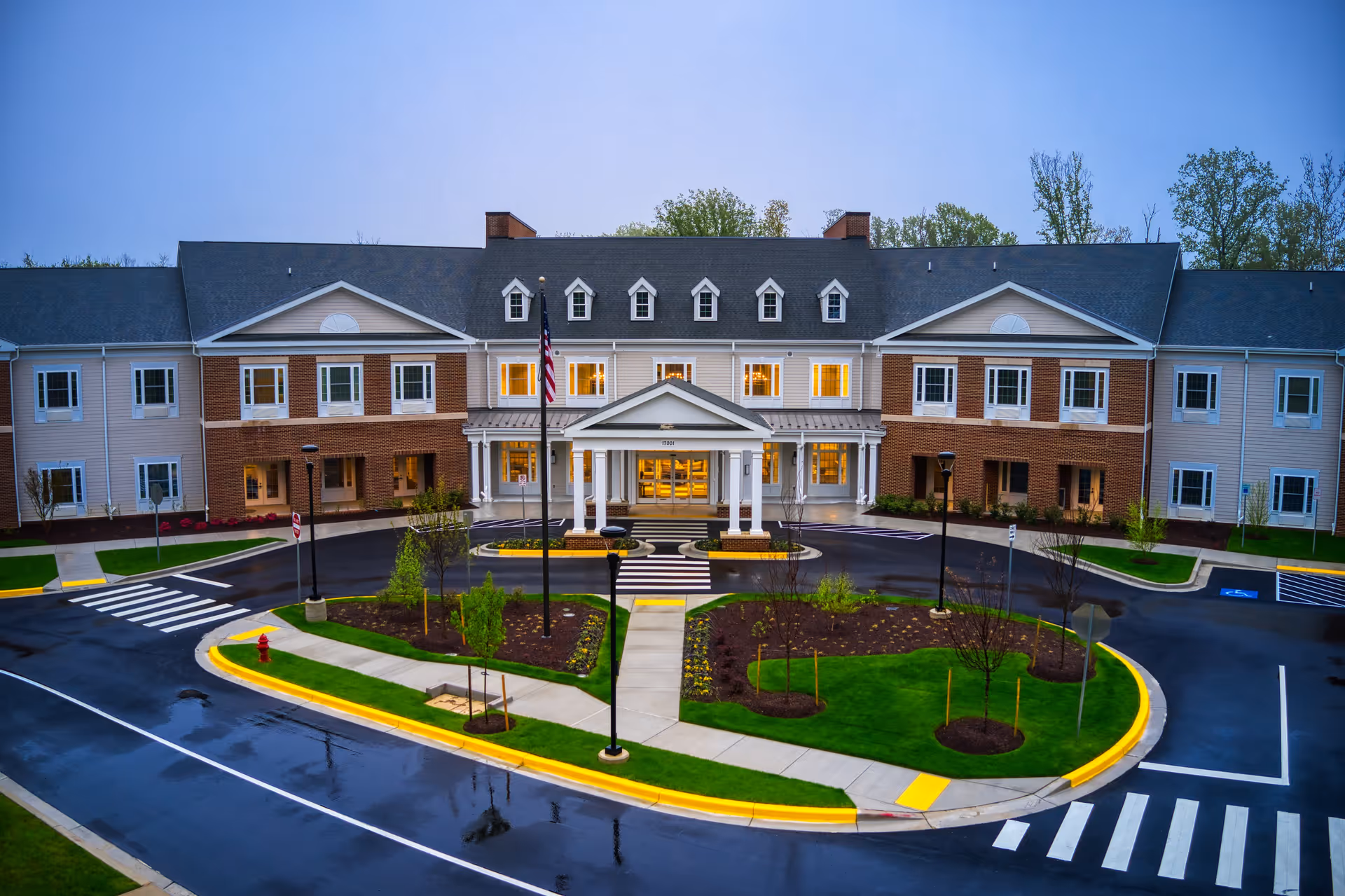 Front exterior of a two-story senior living facility with a covered entrance, circular drive, landscaped island, and an American flag.