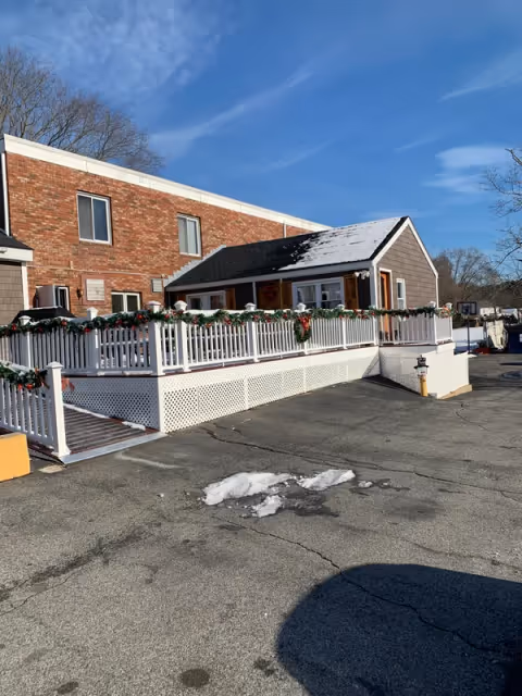 Exterior view of a senior living facility building with a brick upper section and a gray lower section. The building has a white railing decorated with green garlands and red bows, and there is a small amount of snow on the roof and ground. The sky is clear and blue.