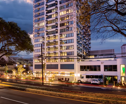 Tall modern high-rise building with lit windows and street-level storefronts at dusk.
