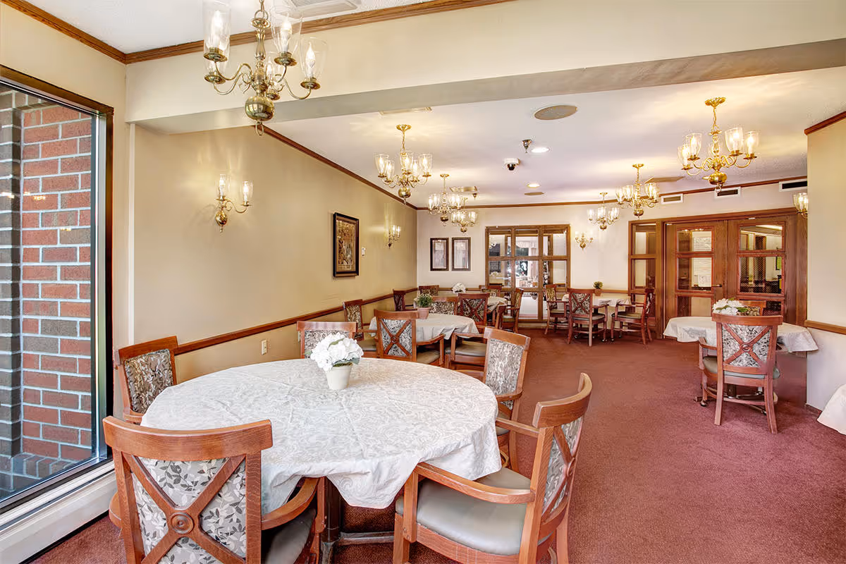 A dining room with round and rectangular tables covered with white tablecloths, surrounded by wooden chairs with patterned cushions. The room features multiple brass chandeliers and wall sconces with glass shades, beige walls with wooden trim, and a large window showing a brick exterior wall.