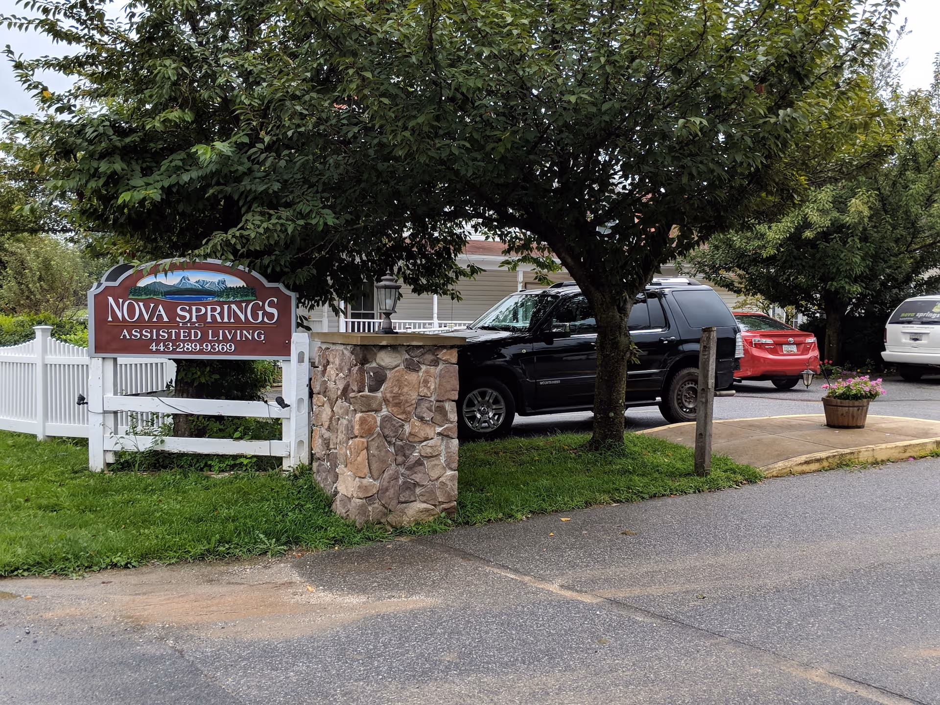 Exterior view of Nova Springs Assisted Living with a sign, stone pillar, trees, and parked cars.