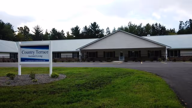 Exterior view of Country Terrace Assisted Living facility showing a single-story building with a metal roof, surrounded by trees and a grassy lawn. A sign in front reads 'Country Terrace Assisted Living' with a website URL below it.
