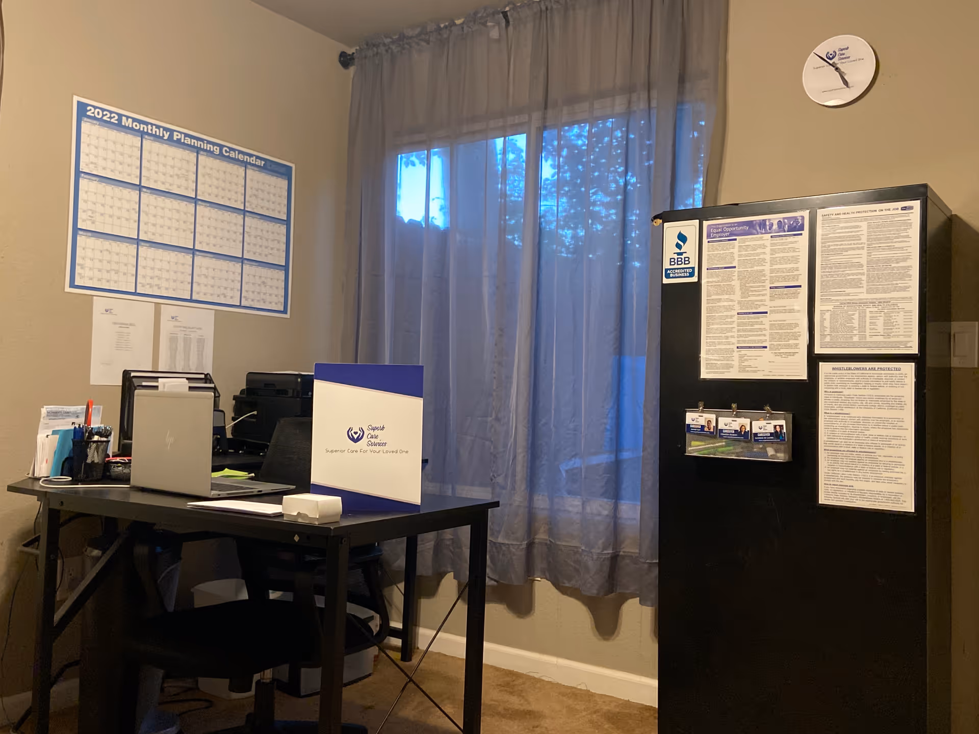 Office desk with laptop, calendar, papers, and a tall black cabinet near a window.