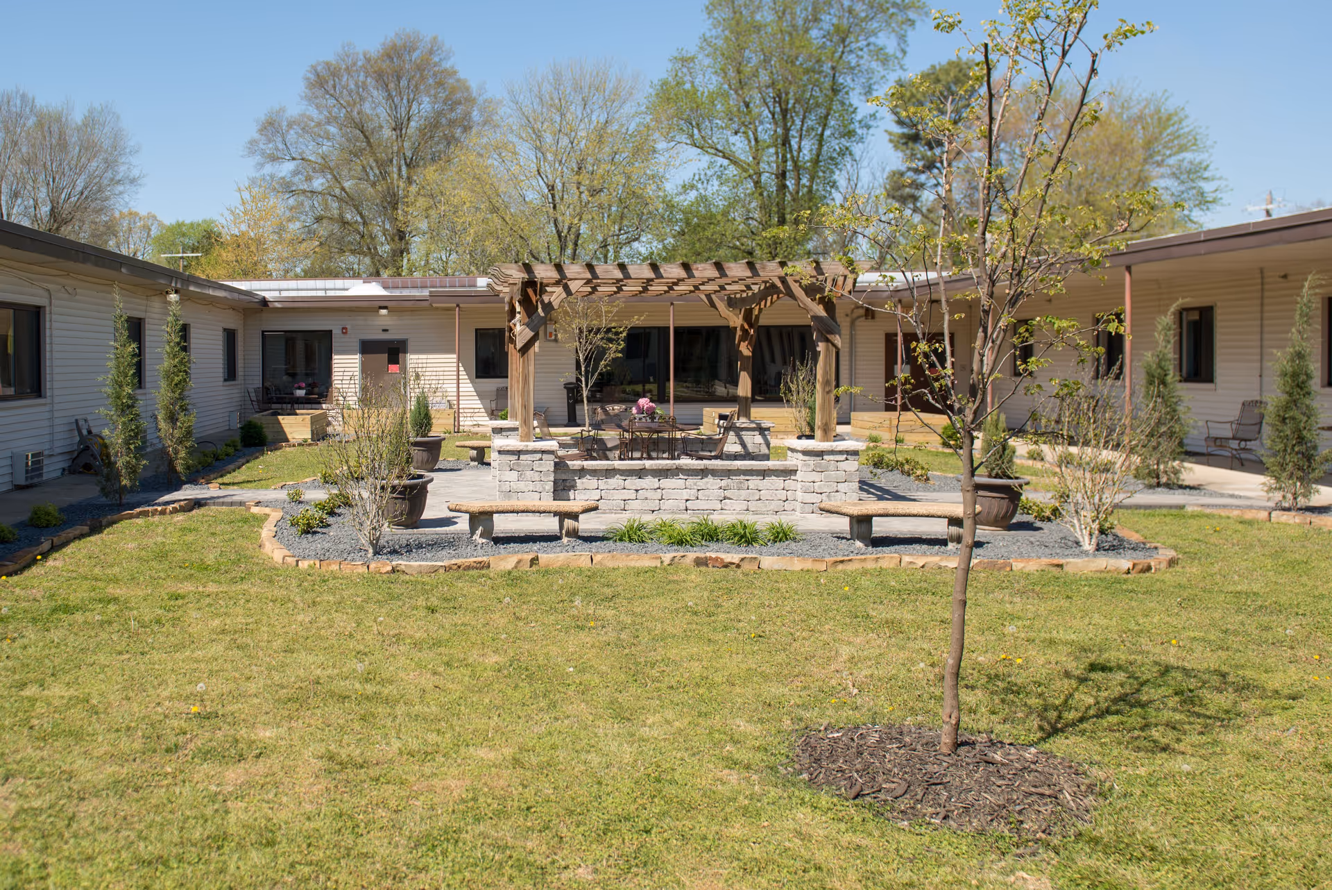 Outdoor courtyard at a senior living center featuring a pergola, seating, planters, and surrounding buildings.