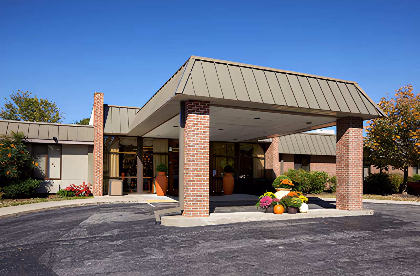 Exterior view of Ahc West Tennessee Transitional Care facility entrance with a covered drop-off area supported by brick columns. There are large windows and doors behind the entrance, with potted plants and colorful flowers arranged near the entrance. The sky is clear and blue, and there are trees and shrubs around the building.
