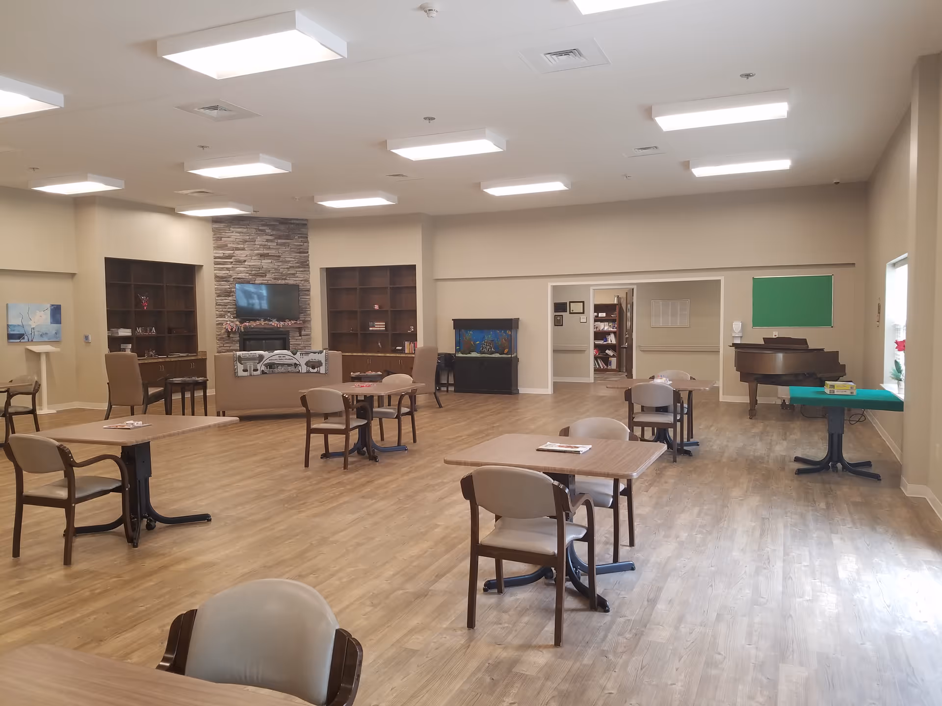 A spacious senior living facility common area with tables, chairs, a piano, and shelving.