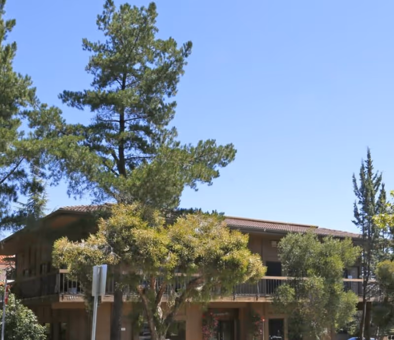 Senior living facility building exterior with balconies, surrounded by tall trees and clear sky.