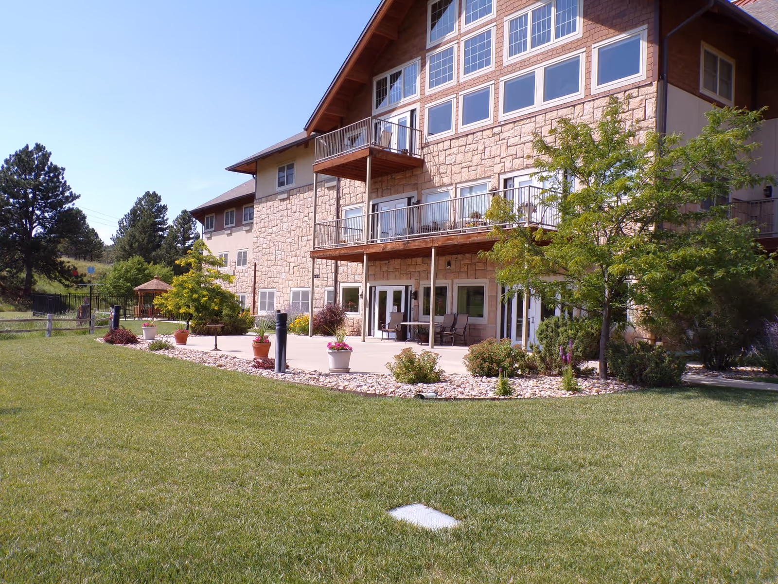 Stone-clad multi-story senior living building with balconies, a patio area, and a well-kept lawn and landscaping.