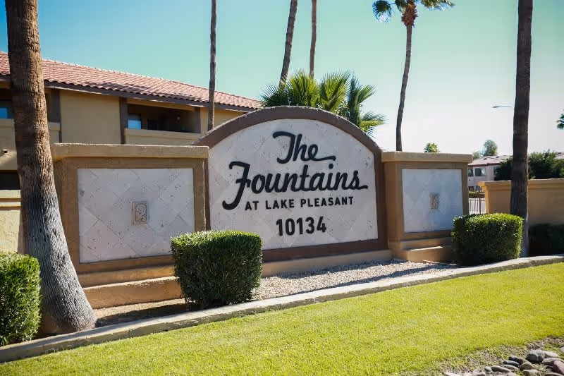 Outdoor entrance sign for The Fountains at Lake Pleasant with the address number 10134, surrounded by palm trees, bushes, and a well-maintained lawn under a clear sky.