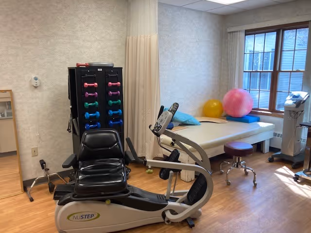 A physical therapy room with exercise equipment including a NuStep recumbent cross trainer, a rack of colorful dumbbells, a treatment table with pillows, exercise balls, a stool, and medical equipment near a window with natural light.