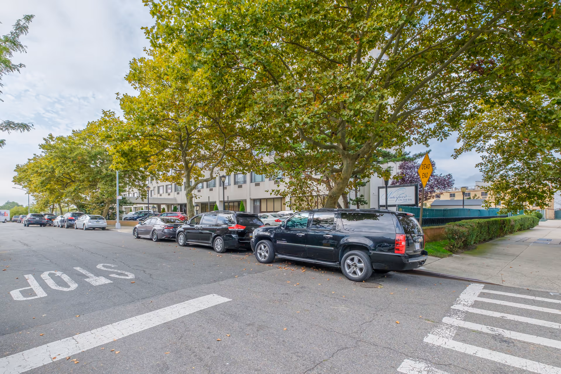Street view of The Waterford on the Bay senior living building with parked cars along a tree-lined curb and a painted STOP on the road.