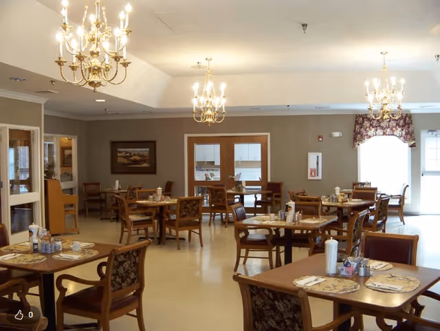 A well-lit dining room with multiple wooden tables and chairs arranged neatly. Each table is set with placemats, napkins, and condiments. The room features beige walls, three ornate chandeliers hanging from the ceiling, a window with floral curtains, and framed artwork on the wall.