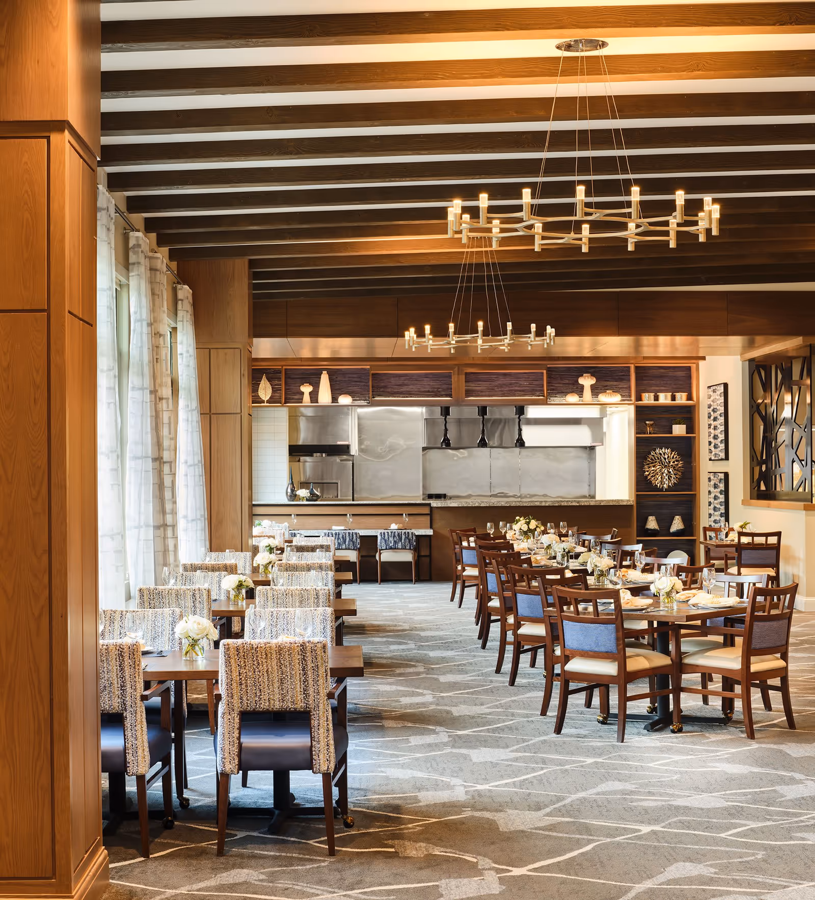 A spacious, well-lit dining room with multiple wooden tables and chairs arranged neatly. The tables are set with glassware, plates, and floral centerpieces. The room features large windows with sheer curtains on the left, wooden beams on the ceiling, modern chandeliers, and an open kitchen area in the background with stainless steel appliances and decorative shelves.