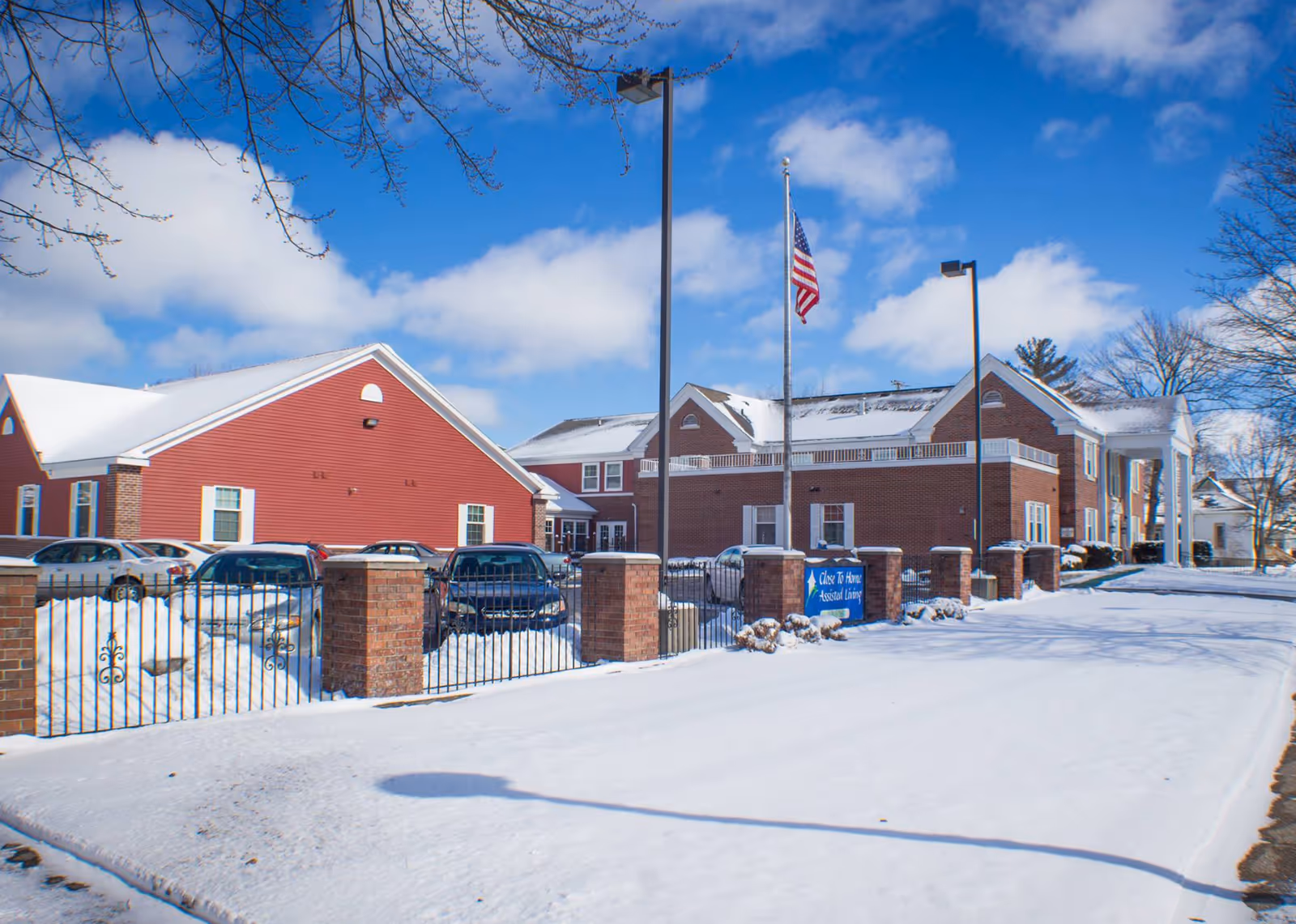 Snow-covered front exterior of a red-brick assisted living building with parked cars, a flagpole, and a fenced entrance.