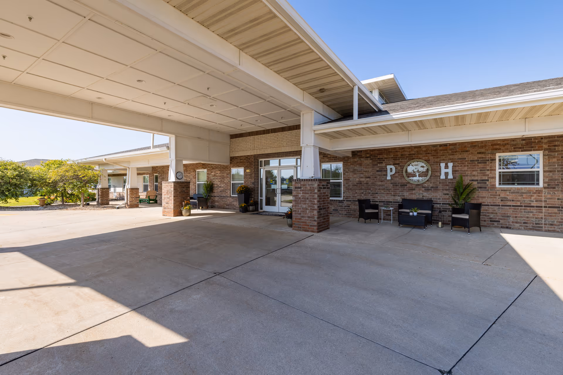 Covered drop-off driveway and main entrance of a brick senior living building with outdoor seating and decorative 'P H' wall signage.