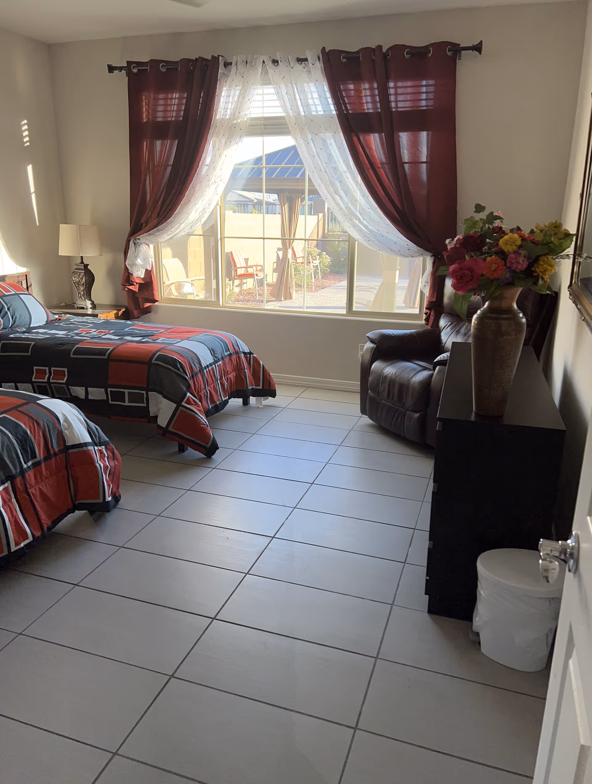 Sunny bedroom with two twin beds, tiled floor, a large window with burgundy curtains, a leather recliner and a dresser topped with a vase of flowers.