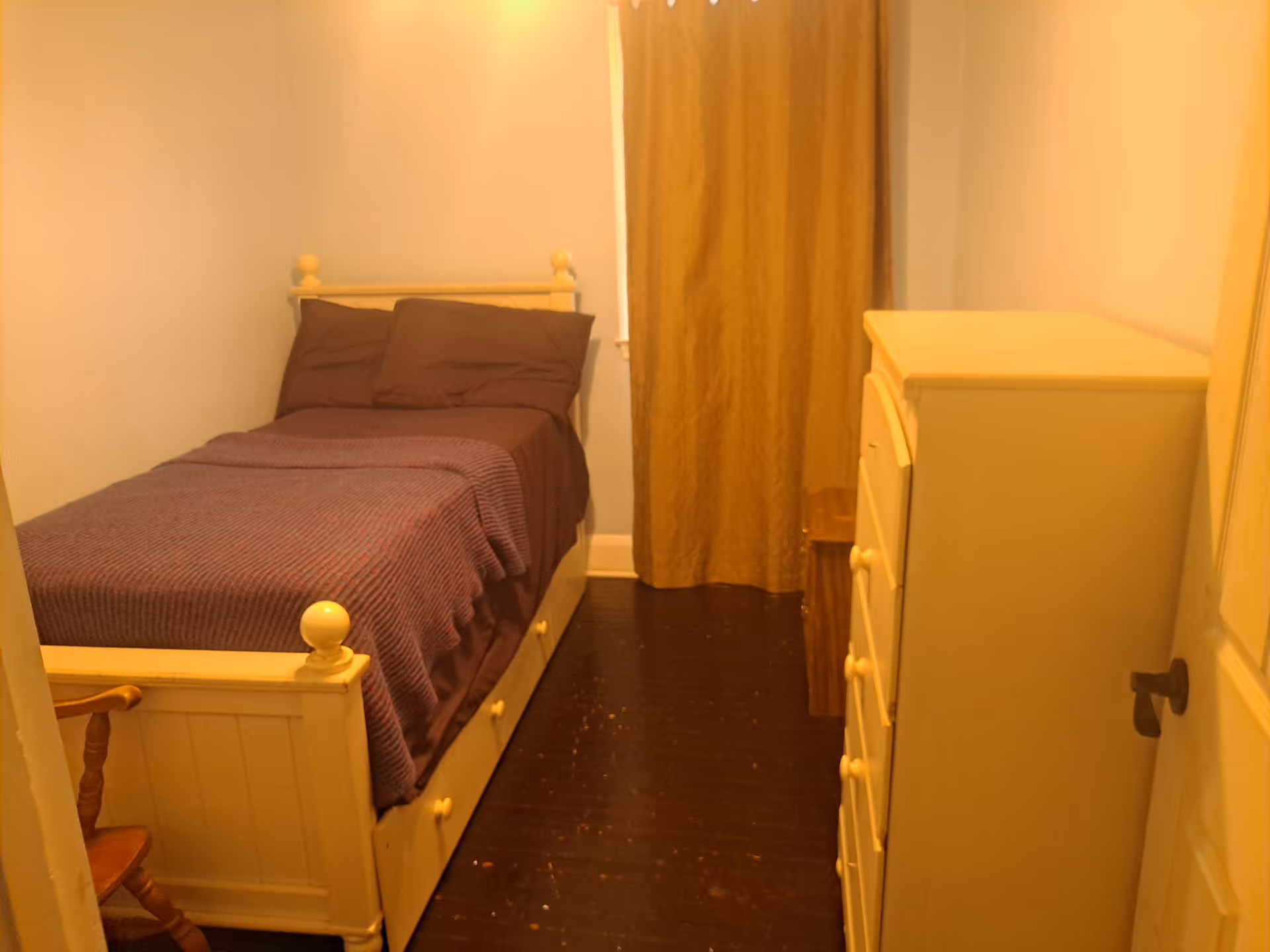 Small bedroom in a senior facility featuring a single bed, dresser, and curtained window.