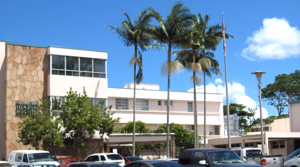 Exterior view of The Queen’s Medical Center Wahiawā with palm trees and parked cars.