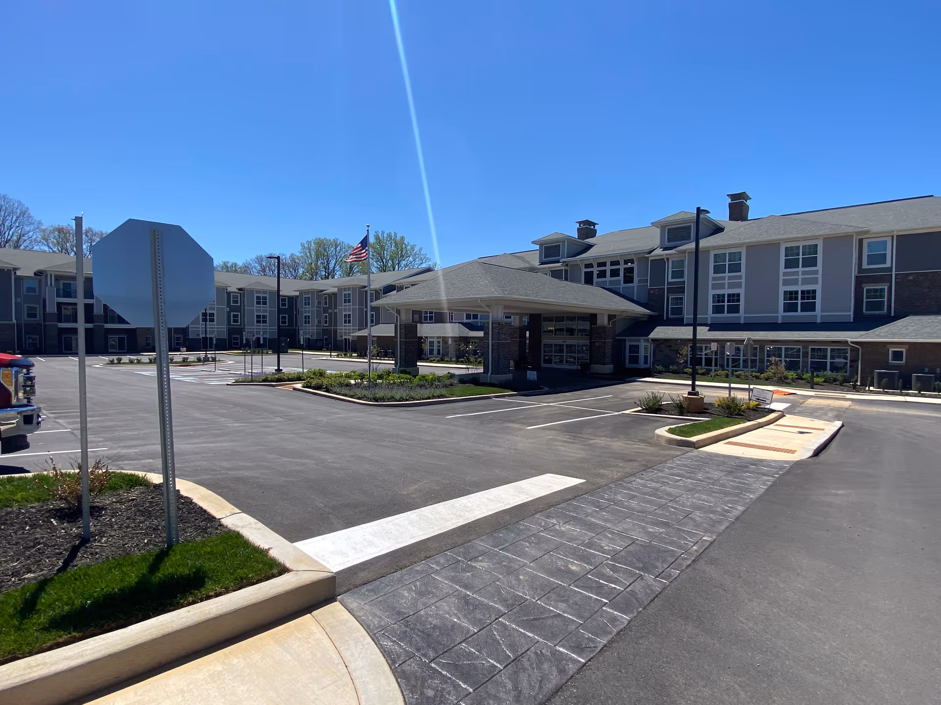 Exterior view of Julian Woods Retirement Community building on a clear sunny day, showing the main entrance with a covered drop-off area, parking spaces, a stop sign, and an American flag on a flagpole.