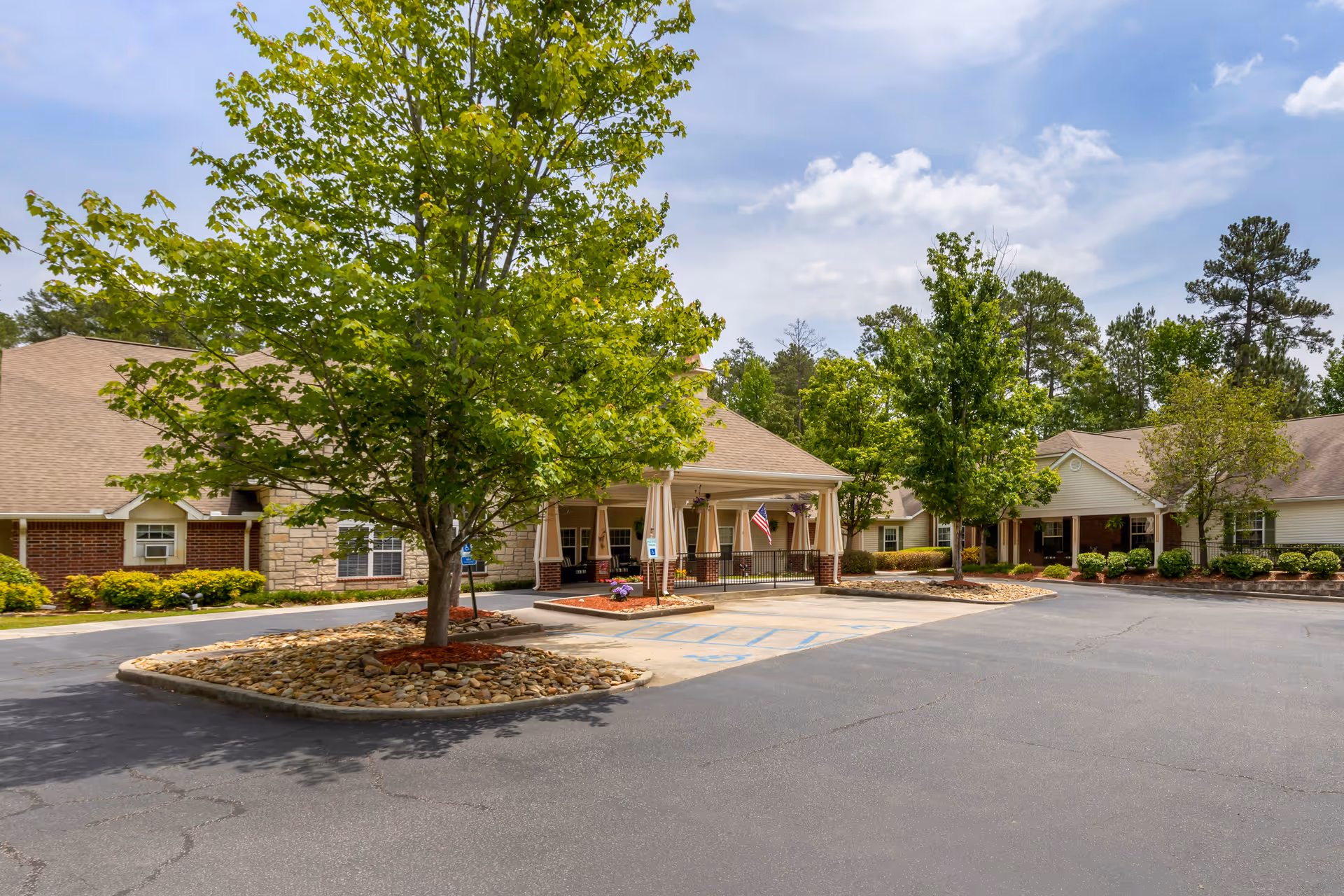 Exterior view of Brookdale Harbison senior living facility showing a driveway with a covered entrance, surrounded by trees and landscaped areas with bushes and rocks under a partly cloudy sky.