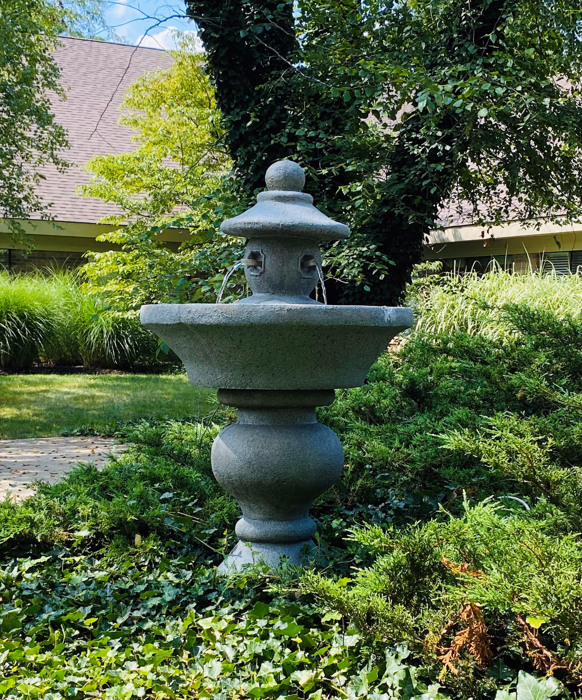 Stone fountain in a landscaped outdoor garden area with greenery and a building in the background.