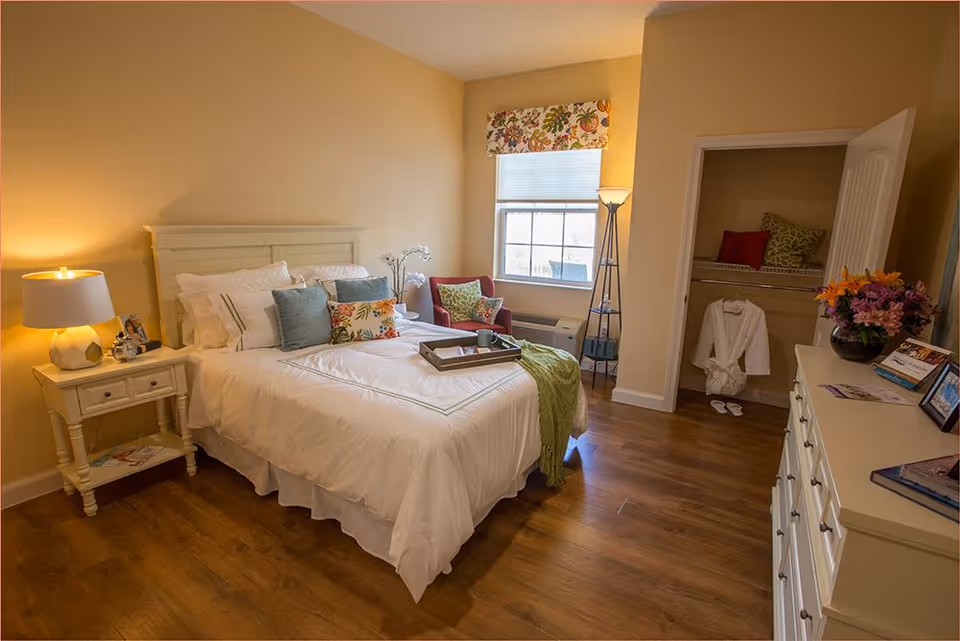 A cozy bedroom with a neatly made bed featuring white bedding and multiple decorative pillows. A wooden nightstand with a lamp and framed photos is beside the bed. There is a window with a floral valance, a red armchair with a green pillow, and a tall floor lamp near the window. An open closet shows a hanging white robe and slippers. A dresser with flowers, photos, and brochures is on the right side of the room. The floor is wooden, and the walls are painted a warm beige color.