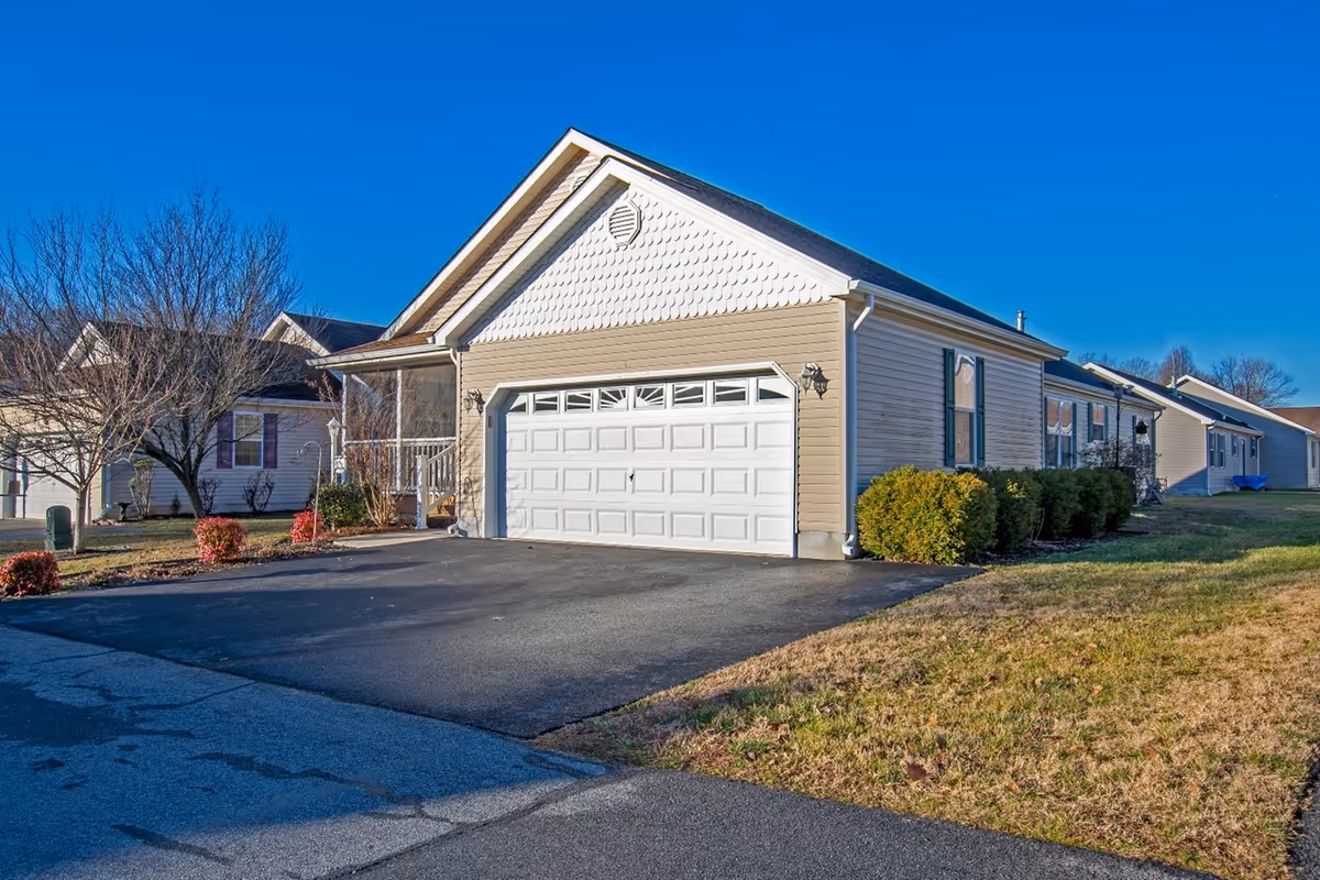 Exterior view of a single-story residential building with beige siding, a white garage door, and a small front porch. The driveway is paved and there are bushes and leafless trees around the property under a clear blue sky.