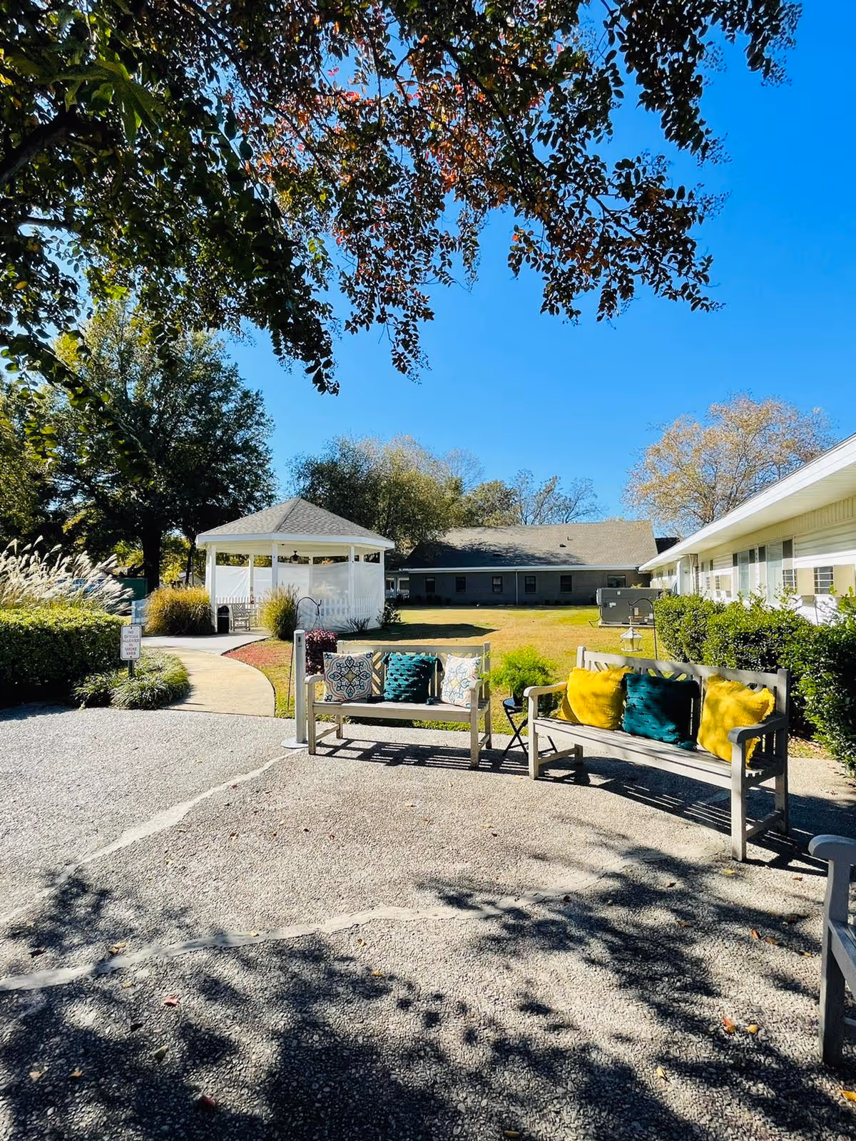 Outdoor seating area at Indianola Rehabilitation and Healthcare Center featuring two wooden benches with colorful cushions, a small side table, a paved pathway leading to a white gazebo, surrounded by green bushes and trees under a clear blue sky.