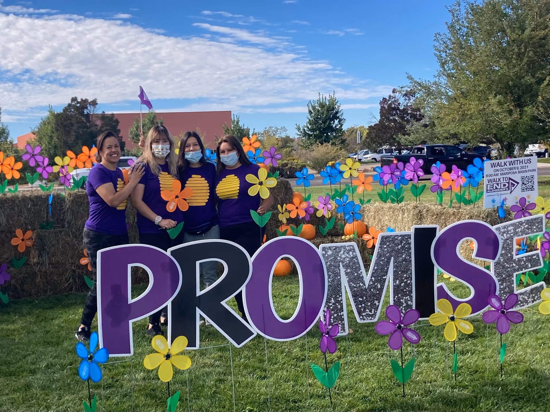 Four women wearing purple shirts with a yellow heart design stand together outdoors on grass, surrounded by colorful flower decorations and hay bales. Behind them is a large sign spelling PROMISE in purple and silver letters. The sky is partly cloudy and there are trees and parked cars in the background.