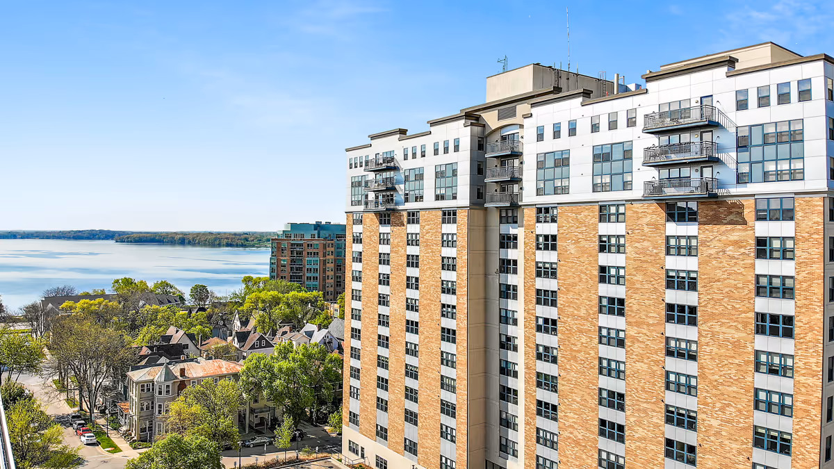 Tall brick and white senior living building with balconies overlooking a lakefront neighborhood.