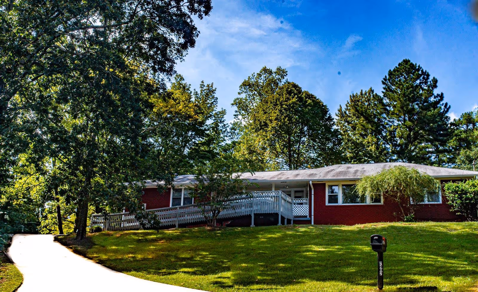Single-story red brick building with a white ramp leading to the entrance, surrounded by green grass and tall trees under a blue sky.