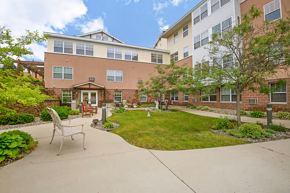 Courtyard of a multi-story senior living building with chairs, walkways, lawn, and landscaping.