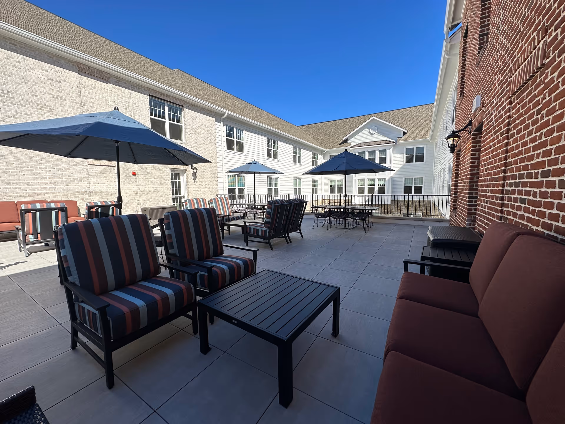 Outdoor patio area at Village Crossing at Worman's Mill with multiple seating arrangements including striped cushioned chairs, a maroon cushioned sofa, black tables, and blue umbrellas under a clear blue sky. The patio is surrounded by a two-story building with white and brick exterior walls.