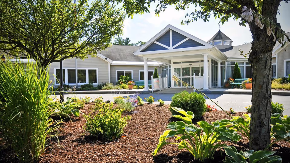 Front exterior view of Havenwood Heritage Heights facility with a landscaped garden in the foreground, featuring various green plants and flowers, and a covered entrance with white columns under a blue sky.