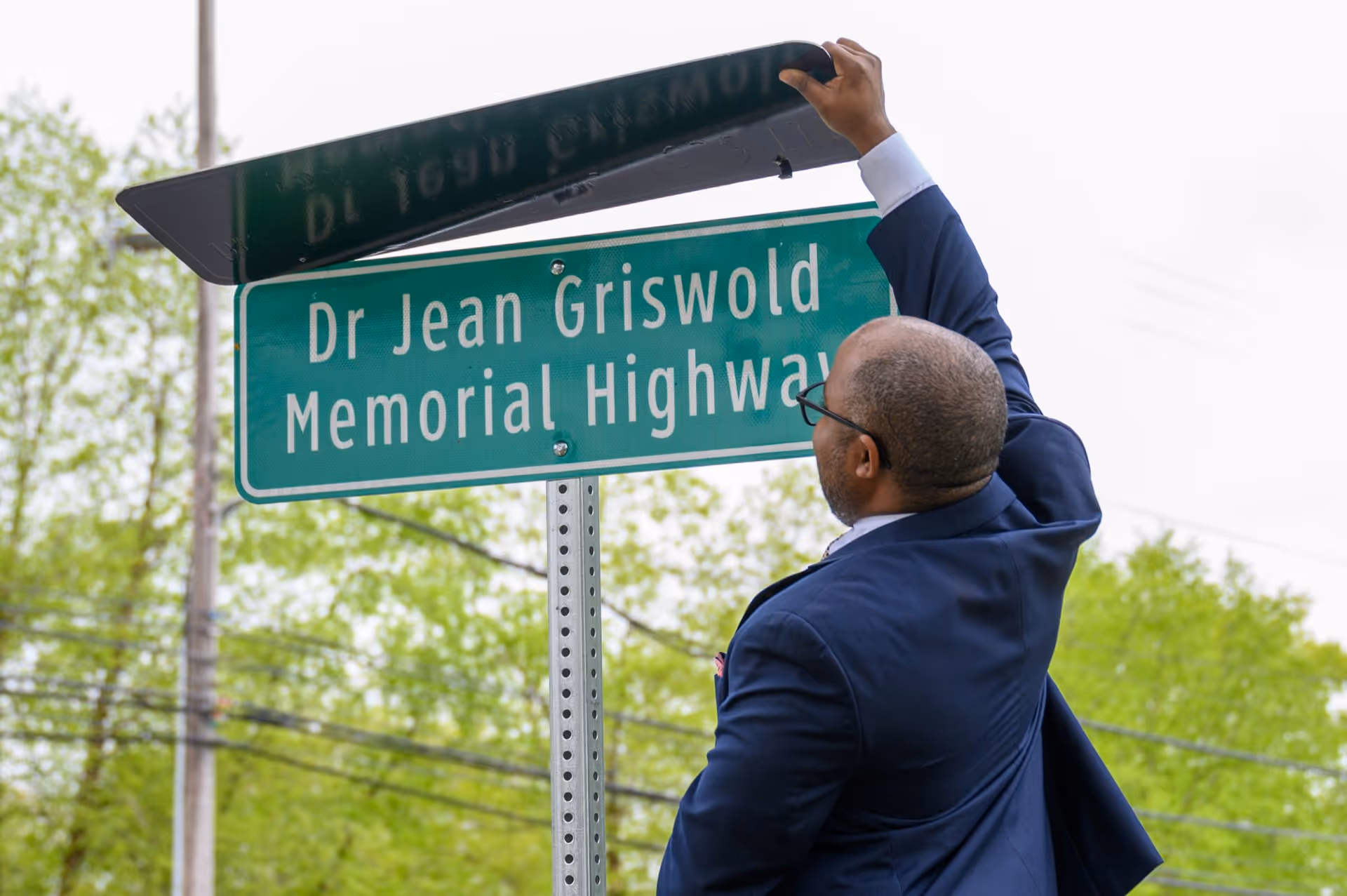 Man in a dark suit adjusts the Dr Jean Griswold Memorial Highway street sign outdoors.