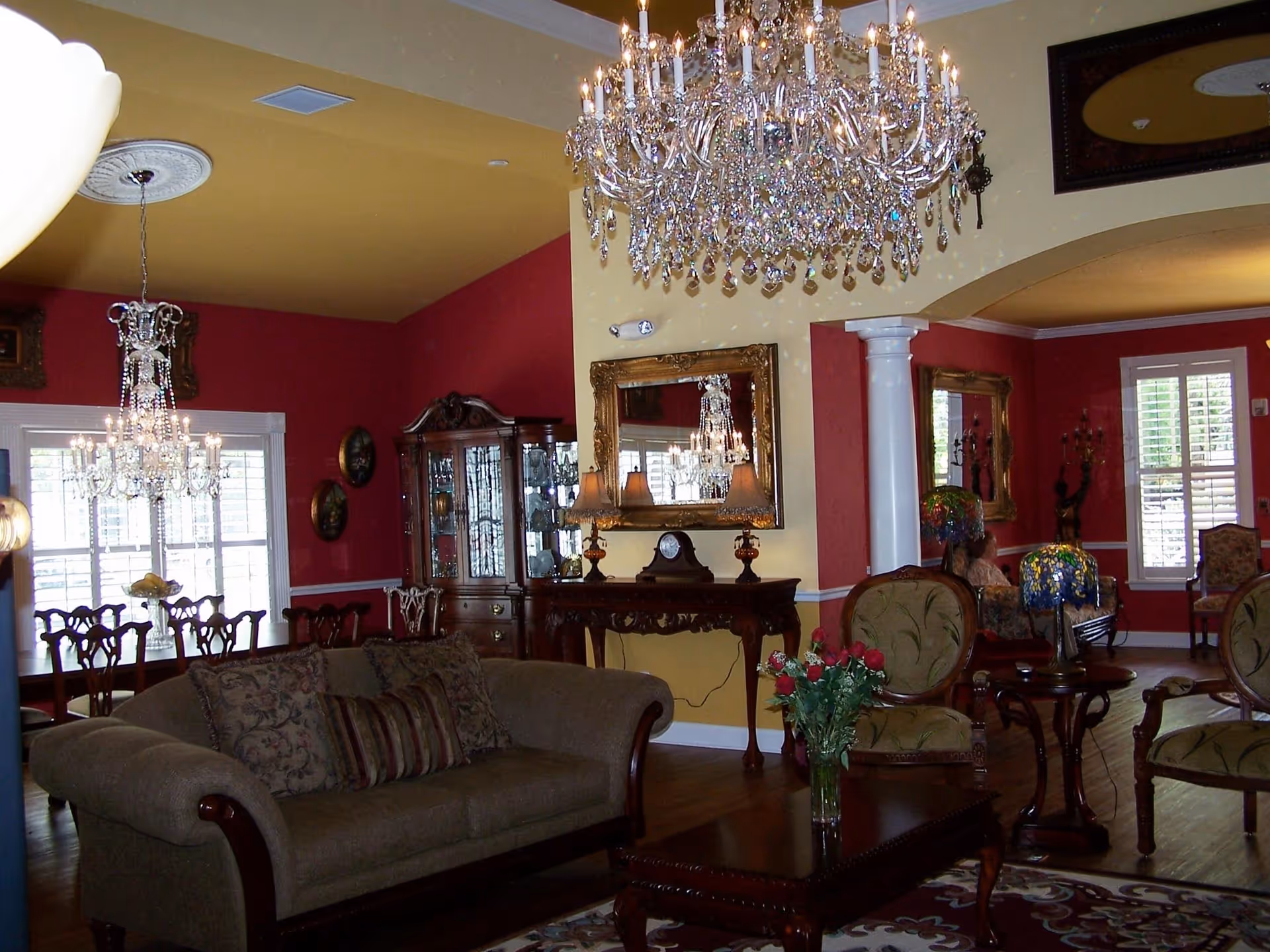 A spacious and elegantly decorated living room with a large chandelier hanging from the ceiling. The room features a beige sofa with patterned cushions, a wooden coffee table with a vase of red roses, and several upholstered chairs. The walls are painted in warm tones of red and yellow, with large mirrors and framed artwork. In the background, a dining area with a chandelier and wooden dining chairs is visible, along with a glass-fronted cabinet displaying decorative items.