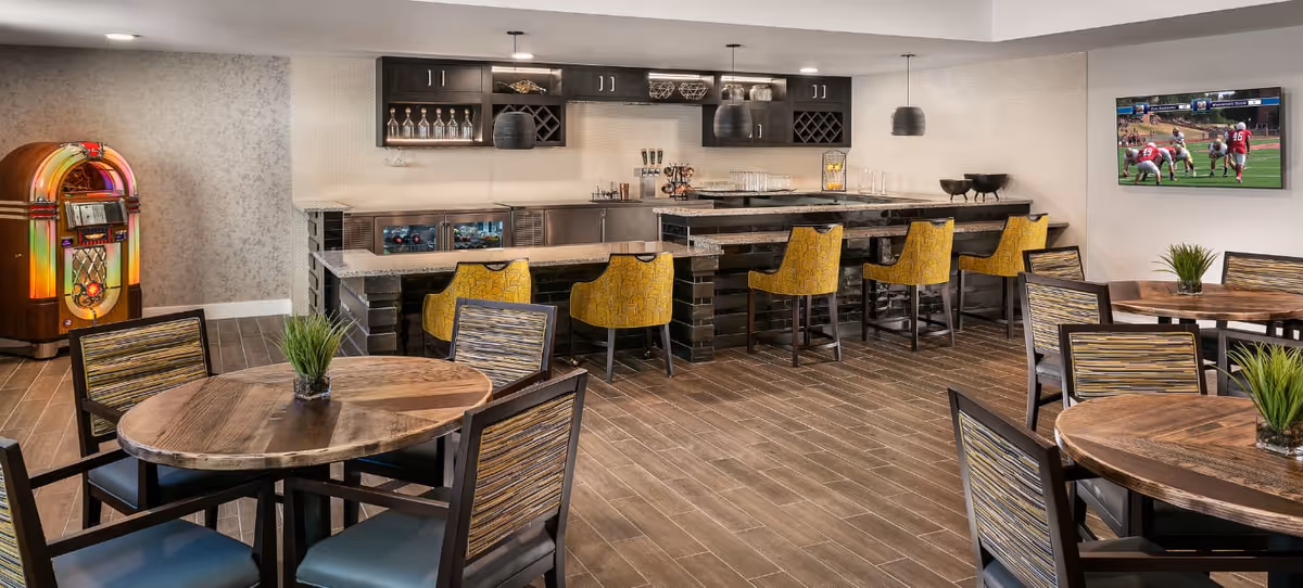 A modern dining area featuring round wooden tables with chairs, a bar counter with yellow upholstered bar stools, a jukebox in the corner, and a wall-mounted TV showing a football game. The space has wood-look tile flooring and dark cabinetry with glassware and decorative items.