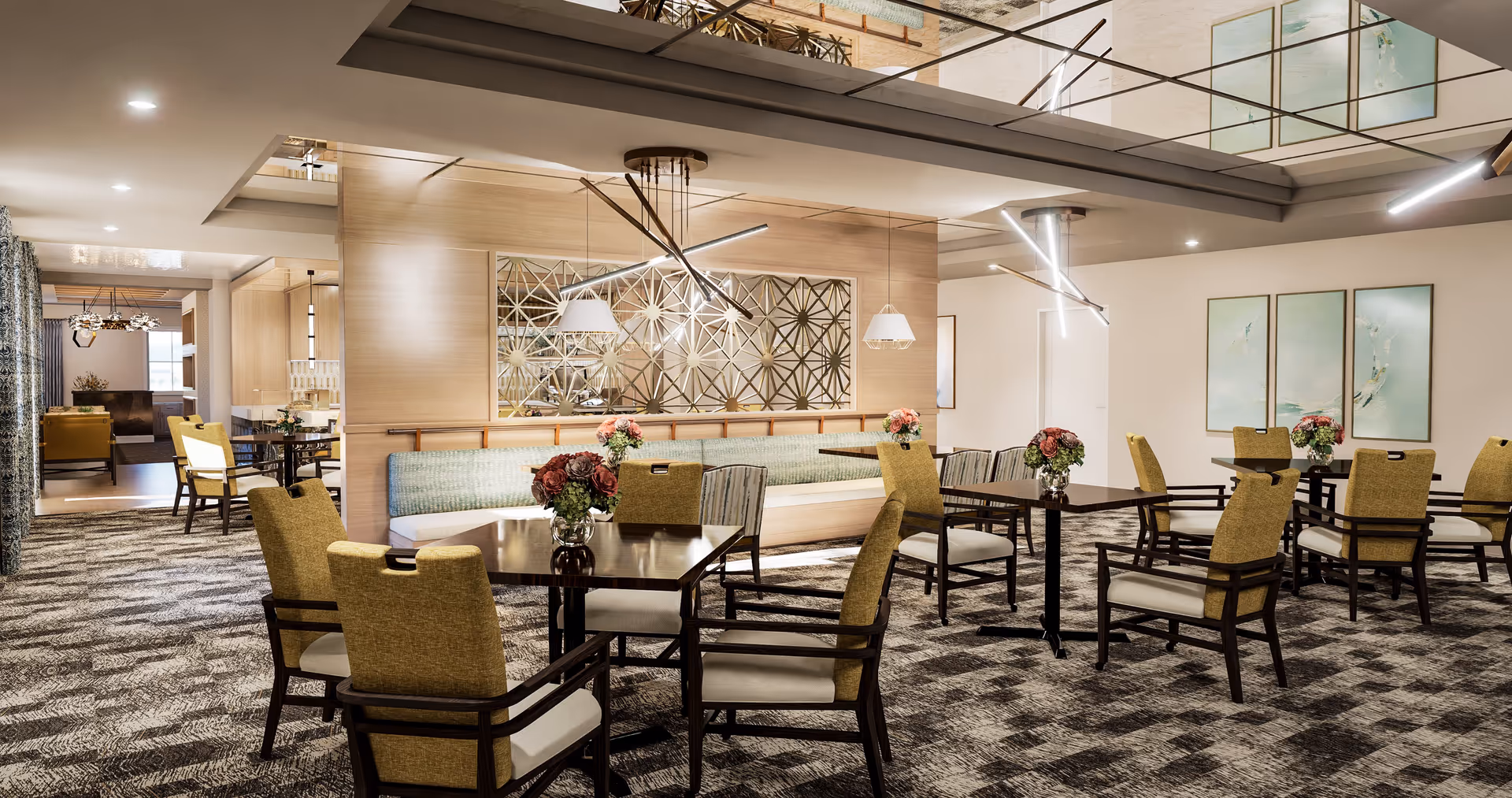 Spacious, well-lit communal dining room with multiple tables and upholstered chairs, pendant lighting, and floral centerpieces.