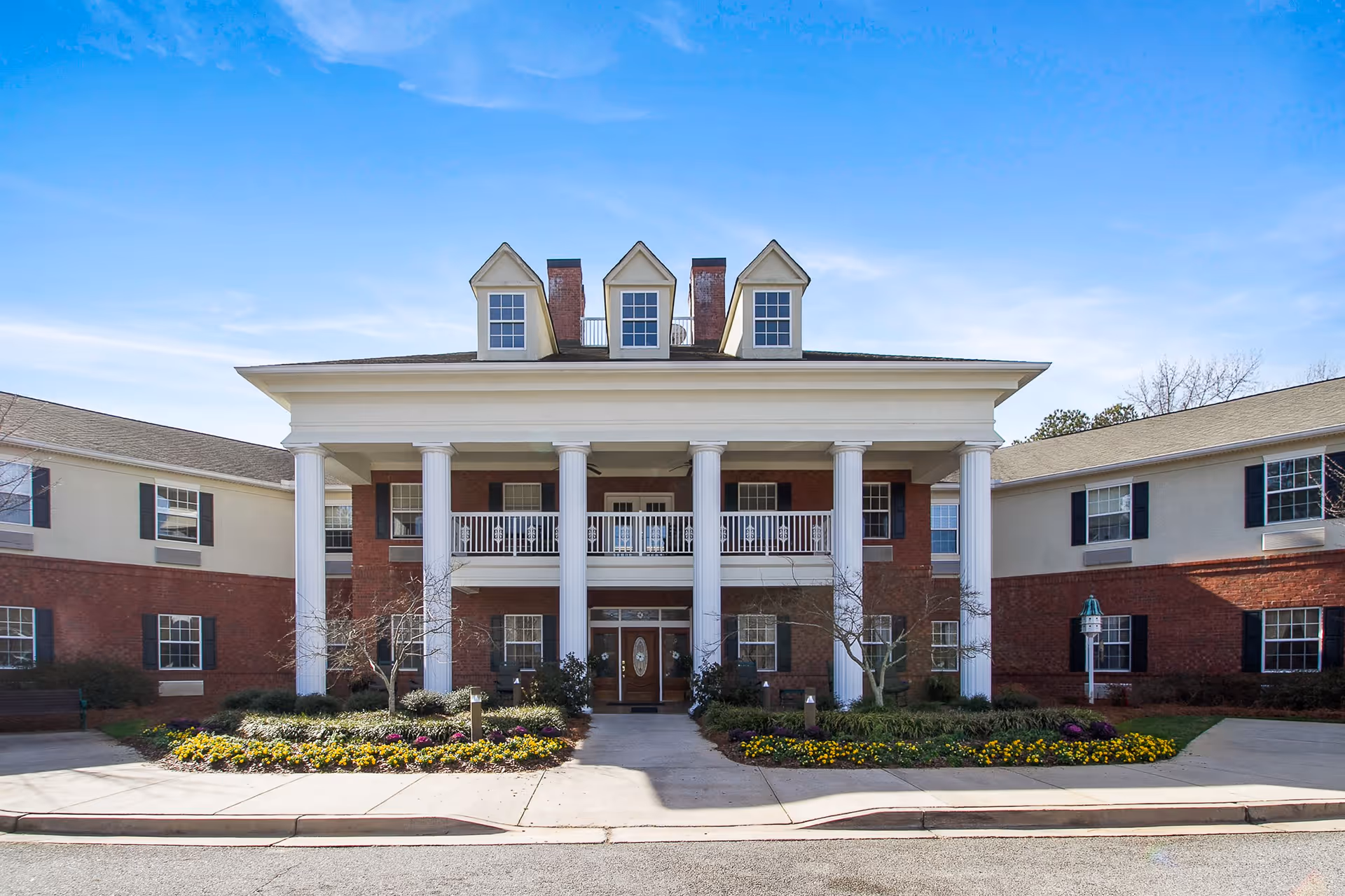 Front exterior view of a two-story senior living facility building with large white columns, a balcony, and three dormer windows on the roof. The building has a brick and beige facade with black shutters on the windows. There are landscaped flower beds and small trees in front of the entrance under a clear blue sky.
