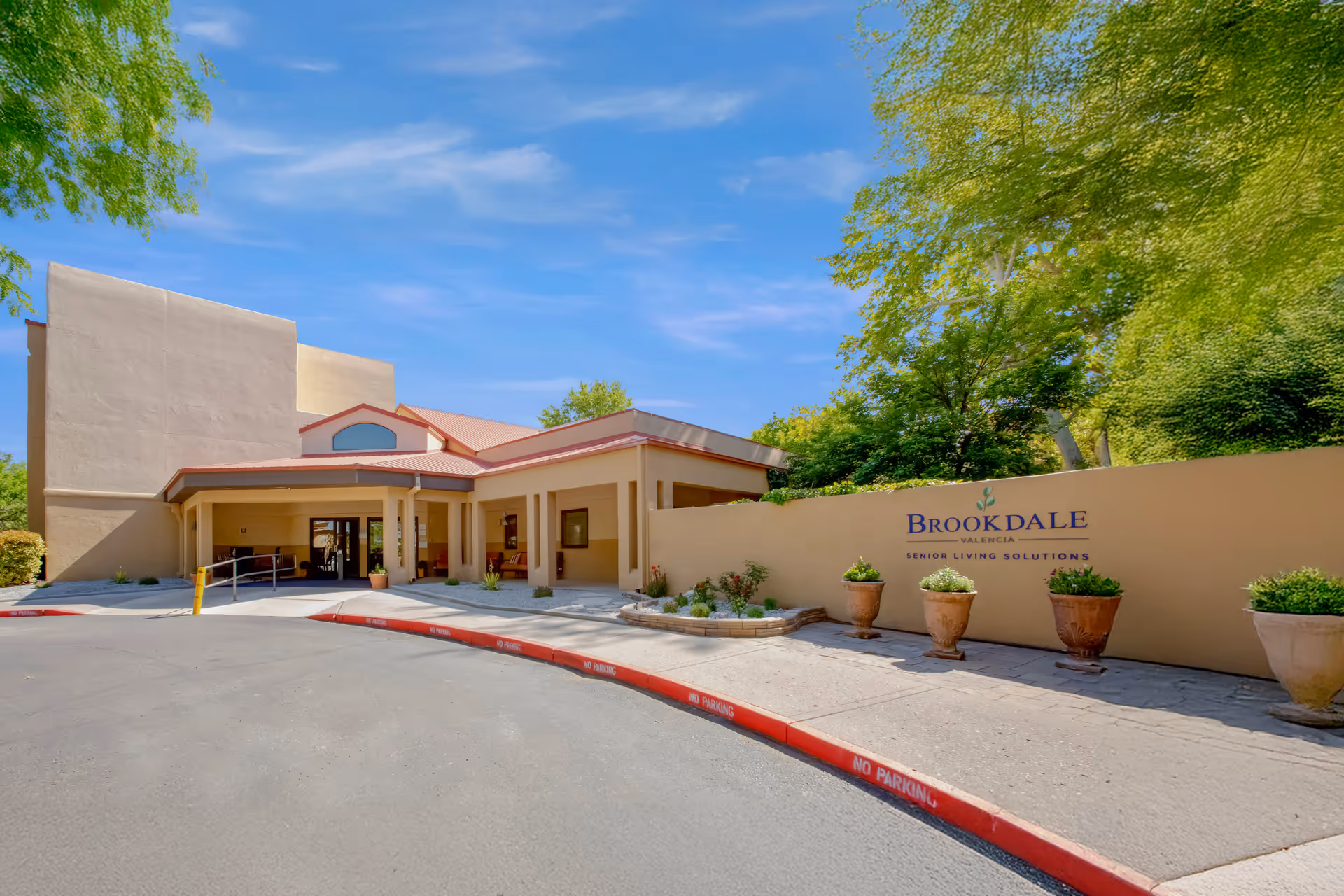 Exterior view of the Brookdale Valencia senior living facility entrance with a beige building, a covered drop-off area, potted plants, and a sign on a wall that reads 'Brookdale Valencia Senior Living Solutions' under a clear blue sky.