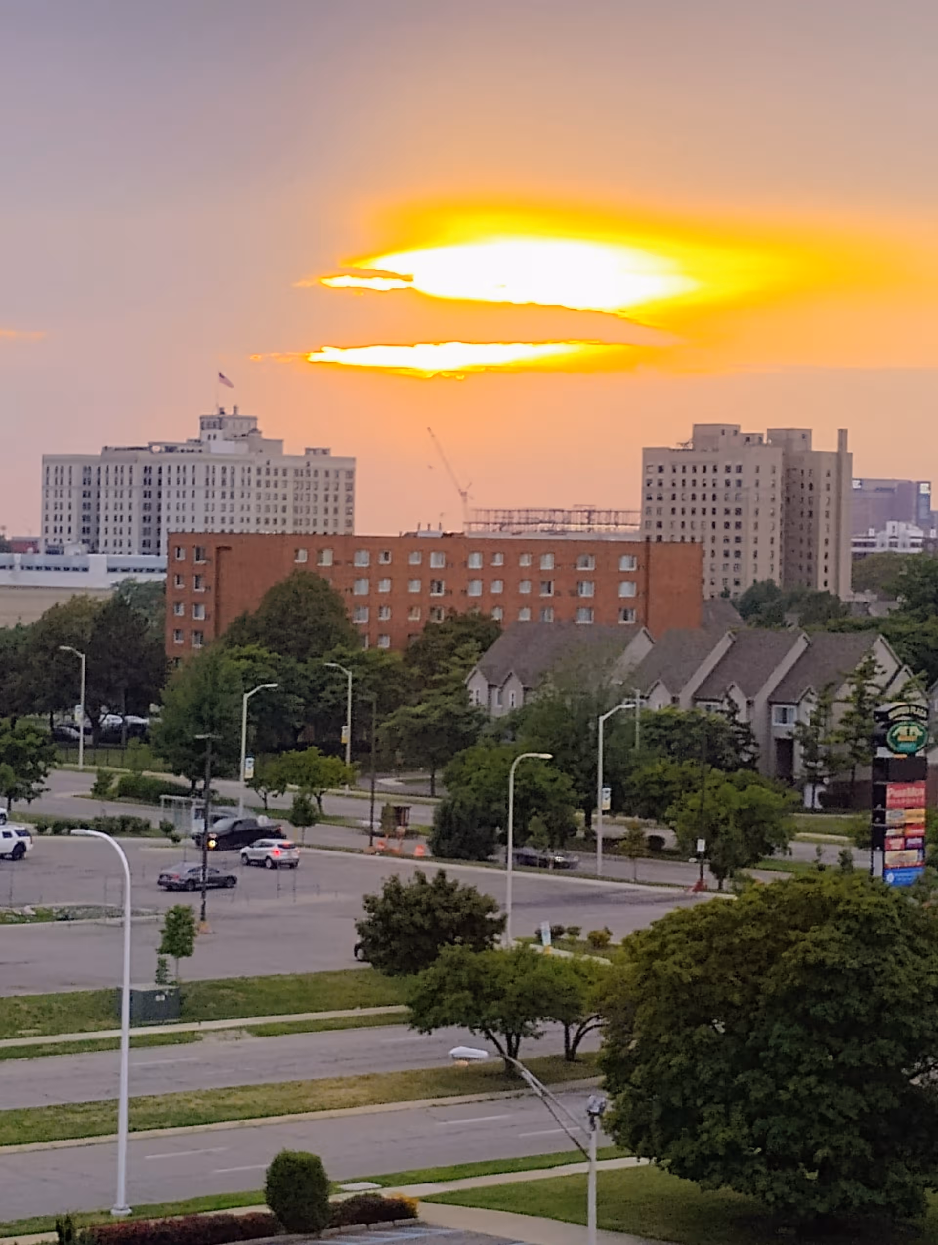 Sunset over a cityscape with multi-story buildings, trees, and a street in the foreground.