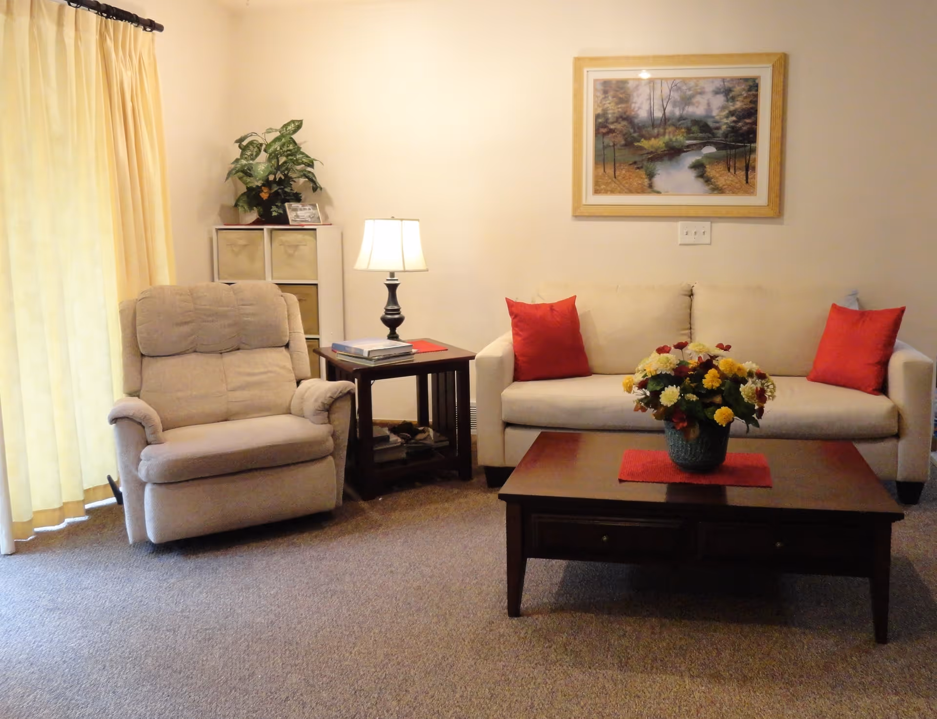 A cozy living room with a beige recliner chair on the left and a beige sofa with two red pillows on the right. Between them is a wooden side table with a lamp and some books. In front of the sofa is a wooden coffee table with a vase of colorful flowers. A framed landscape painting hangs on the wall above the sofa, and yellow curtains cover a window on the left side.