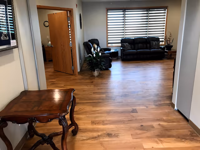 Cozy facility living room with leather sofas, wooden floor, plants, and large window blinds.