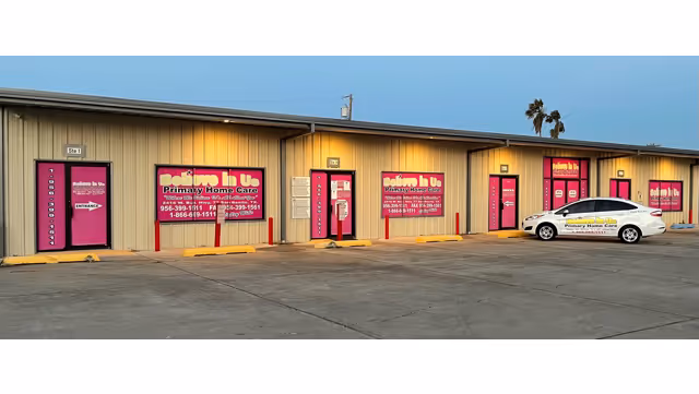 A row of beige storefront units with pink signs advertising Believe In Us Primary Home Care