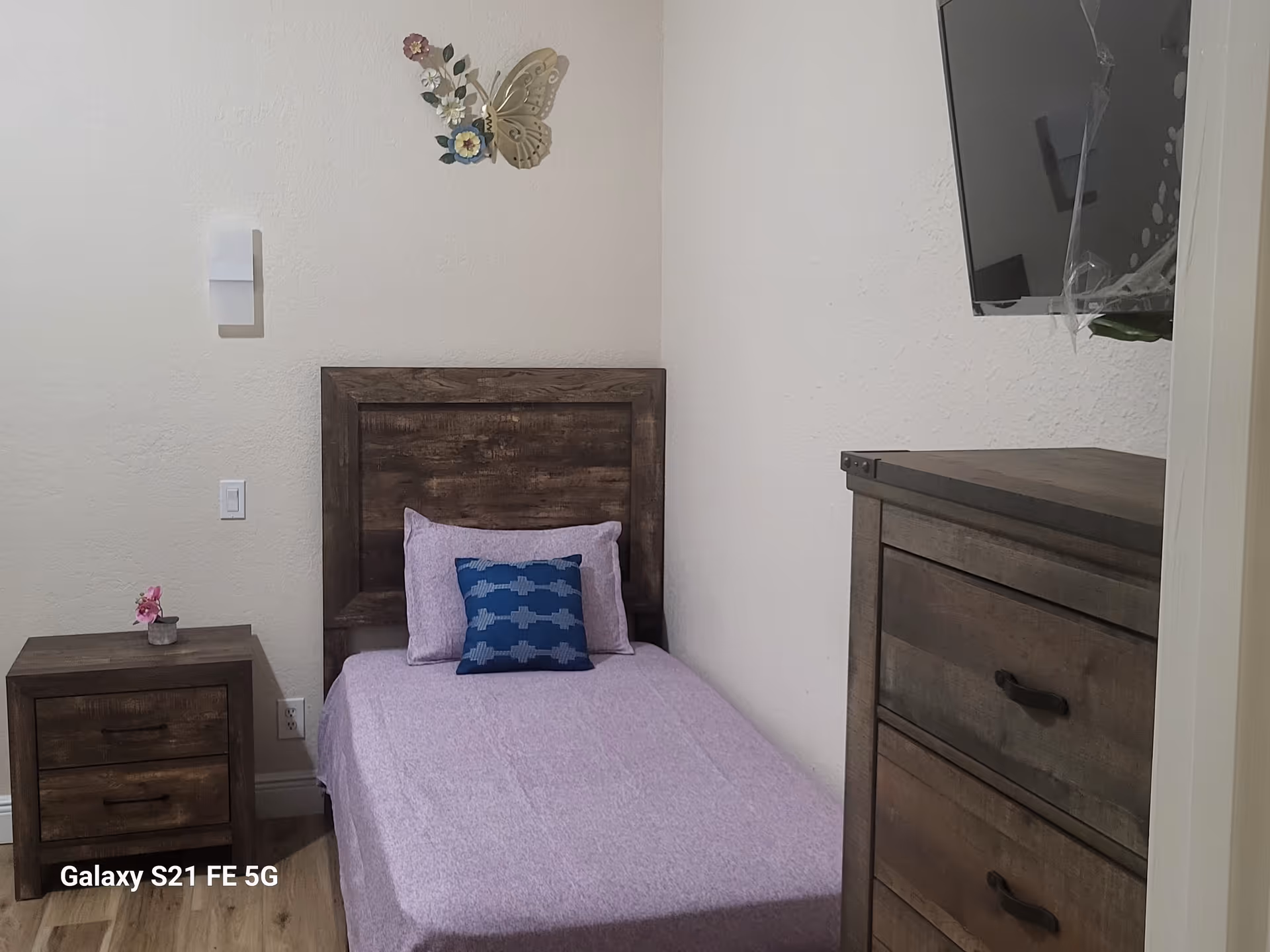 A small bedroom with a wooden bed, lilac bedding, nightstand, dresser, and wall TV.